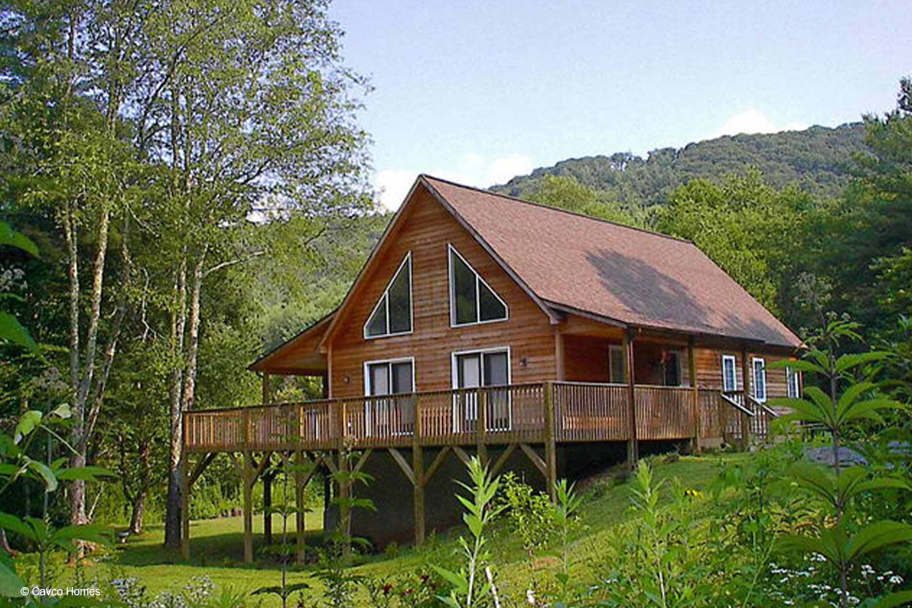 A modular home with large windows sits on a raised deck surrounded by trees and greenery in a peaceful mountain setting.