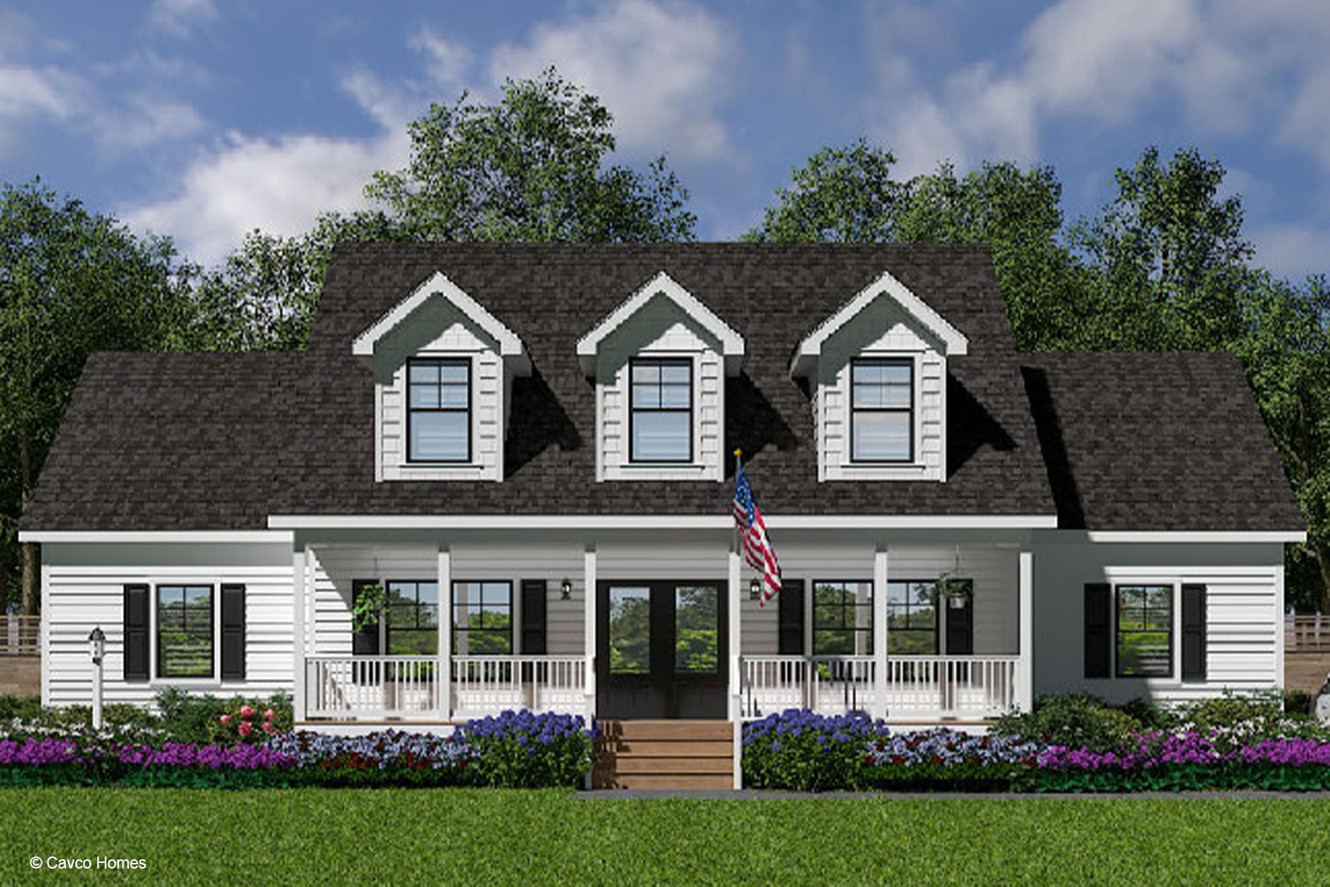 Charming modular home with a black roof, three dormer windows, front porch, American flag, and colorful flowerbeds.